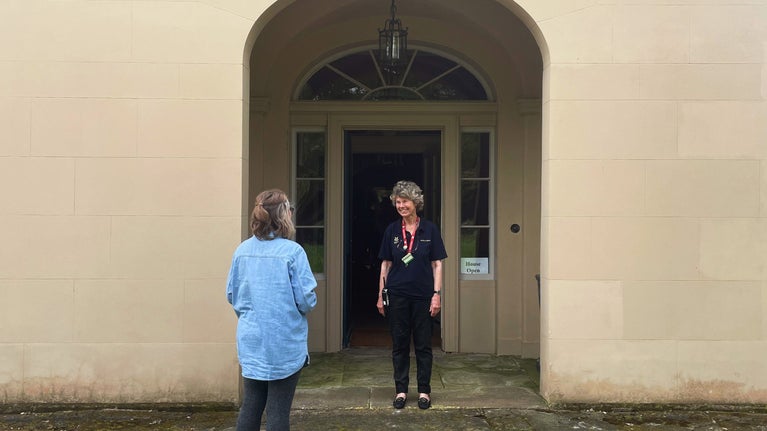 A visitor stands outside the entrance of a historic house, greeted by a smiling staff member. The staff member wears a dark uniform with a lanyard and badges, while the visitor is casually dressed in a light blue shirt and gray pants. The entrance features a large arched doorway with an ornate fanlight window above and a hanging lantern. A sign near the door reads "House Open," indicating that the location is welcoming guests inside.